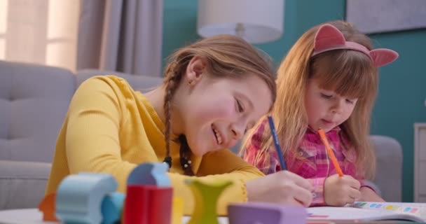 Detailní záběr legračních malých bělošských sester, které jsou samy doma a radostně si spolu hrají. Hezké děti zbarvení obrázek s barevnými tužkami v útulné místnosti. Děti se baví, hrají si a chatují. — Stock video