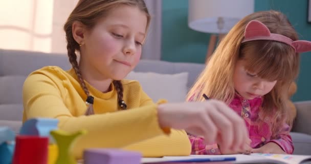 Close up of cheerful small Caucasian sisters sitting at home alone and playing nice together. Cute kids coloring picture with colorful pencils in cozy room. Happy children play and chatting indoor. — Stock Video