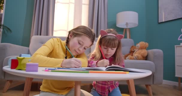 Kavkazské malé roztomilé děti sedí v útulném pěkném pokoji u stolu a zbarvení obrázků s barevnými tužkami. Veselé děti se baví a hrají si uvnitř. Holky zůstávají doma samy. — Stock video