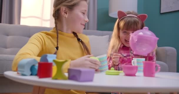 Cute kaukaskie dziewczynki bawiące się zastawą stołową dziecka w przytulnym salonie. Mniejsza siostra nalewa wymyśloną herbatę od czajnika do starszej. Dzieci bawią się razem w domu będąc same. — Wideo stockowe