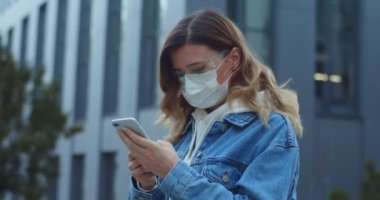 Şehirde maskeli akıllı telefon takan bir kadın. Sağlık ve güvenlik, N1H1 koronavirüs, virüs koruma ve tıbbi konsept. Sokakta koruyucu tıbbi maskeli genç bir kızın portresi..