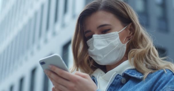 Kvinnan bär mask med smart telefon i stan. Hälsa och säkerhet, N1H1-koronavirus, virusskydd och medicinskt koncept. Porträtt av en ung flicka med skyddande medicinsk mask på gatan. — Stockvideo