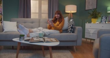 Beyaz kadın evdeki kanepede oturuyor ve web kamerasıyla akıllı telefondan video sohbet ediyor. Güzel, kızıl saçlı bir kadın, oturma odasında konuşuyor ve görüntülü sohbet ediyor..