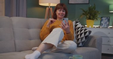 Beyaz tenli güzel kızıl saçlı kadın oturma odasında kanepede oturuyor ve web kamerasıyla akıllı telefonunda video sohbet ediyor. Güzel görünümlü, neşeli kadın konuşuyor ve rahat bir dairede görüntülü sohbet ediyor..