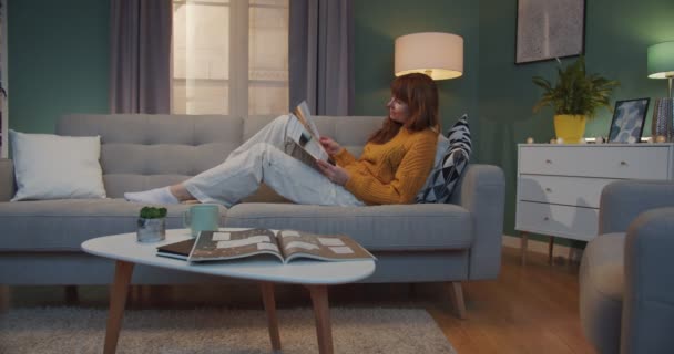 Hermosa mujer de pelo rojo caucásica acostada en un sofá acogedor y leyendo la revista de moda en la sala de estar. Homie mujer descansando en bonito apartamento en fin de semana y volteando páginas de diario perezoso . — Vídeo de stock