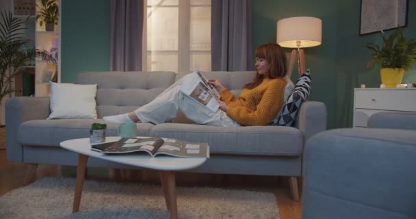 Jolie femme blanche aux cheveux roux couchée sur un canapé et lisant un magazine dans un salon confortable. Femme accueillante dans des lunettes reposant dans un appartement confortable le week-end et flipping pages de journal paresseux . — Video