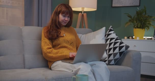 Blanke glimlachte mooie vrouw met rood haar zittend op de bank met laptop computer en rustend in de woonkamer. Vrouwelijk typen op toetsenbord en surfen online tijdens het glimlachen en het weekend thuis doorbrengen. — Stockvideo