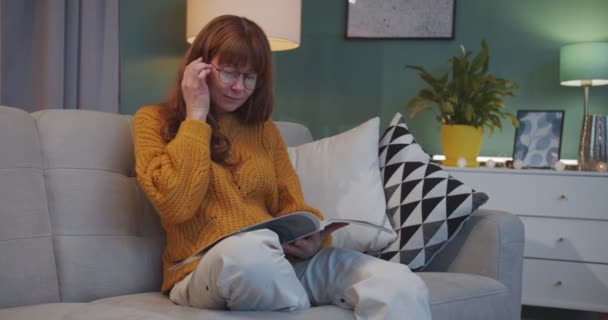 Красивая кавказская рыжая женщина в очках сидит на уютном диване и читает модный журнал в гостиной. Женщина-домохозяйка отдыхает в красивой квартире на выходных и листает страницы журнала ленивый . — стоковое видео