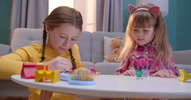 Kaukasische lustige kleine Schwestern, die allein zu Hause Nägel polieren und lackieren. Kleine Kinder spielen im Schönheitssalon im gemütlichen Wohnzimmer. Nette Kinder bei Maniküre drinnen. — Stockvideo