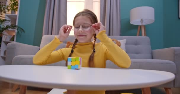 Petite adolescente caucasienne en lunettes jouant avec Ruby Cube à la maison à table. Petit joli enfant qui compose Rubiks cube dans le salon confortable . — Video