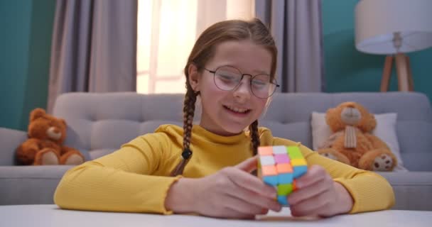 Caucásico niña adolescente en gafas jugando con Ruby Cube en casa en la mesa. Pequeño niño bonito que compone cubo Rubiks en acogedora sala de estar y estar enojado como no puede manejar y hacer frente a ella . — Vídeo de stock