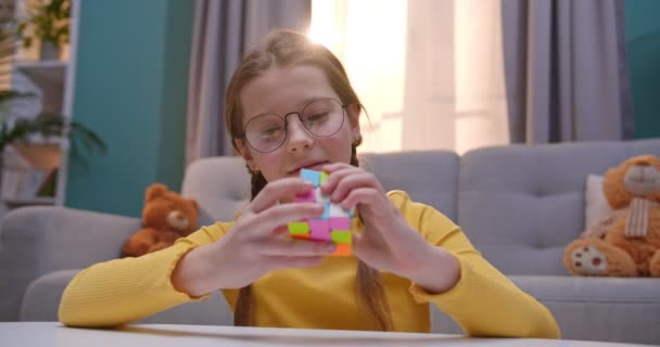 Caucásico niña linda adolescente en gafas jugando con Ruby Cube en casa en la mesa. Bonito niño bonito que compone cubo Rubiks en acogedora sala de estar y no puede hacer frente a ella . — Vídeo de stock