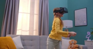 Oturma odasında sanal gerçeklikte oynayan, VR gözlüklü sevimli beyaz kız. Küçük neşeli çocuk, evde dolaşırken kulaklık takıyor. Sanal dünya konsepti.