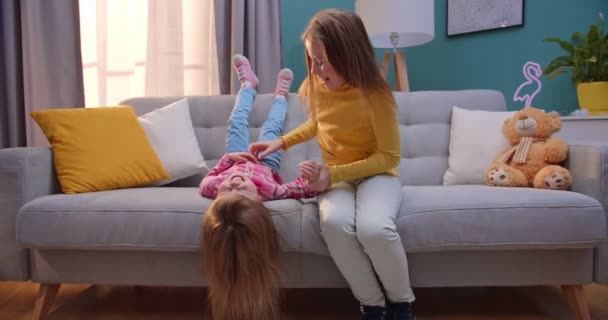 Deux sœurs mignonnes caucasiennes jouant joyeusement sur le canapé dans le salon confortable. Les enfants s'amusent en restant seuls à la maison. Soeur aînée chatouillant plus jeune plus sage ou ami sur le canapé . — Video