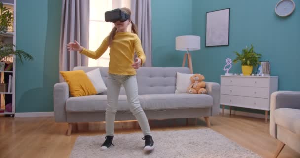 Kaukasisches süßes Teenie-Mädchen mit VR-Brille steht im Wohnzimmer und spielt in virtueller Realität wie Dinge berühren und greifen. Lustiges fröhliches Kind mit Headset drinnen. Konzept der Virtualität. — Stockvideo