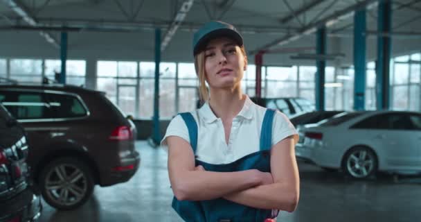 Portrét krásné bělošky auto mechanik v uniformě a klobouk stojící v hale s auty. Hezká dívka se vesele usmívá na kameru v garáži. Koncept údržby automobilu. — Stock video