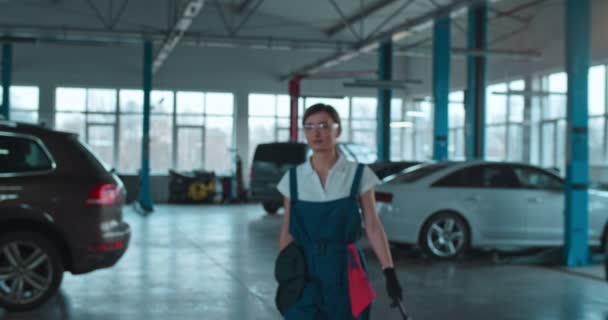 Caucasiano jovem trabalhador de serviço de automóvel feminino em uniforme, óculos e com chave aproximando-se da câmera em grande garagem com carros. Retrato de menina bonita mecânico sorrindo alegremente . — Vídeo de Stock