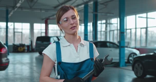 Porträtt av ung vit vacker kvinna auto mekaniker i goggles och med verktyg ler mot kameran och ger tummen upp i garaget. Ung söt flicka i uniform och hatt med skiftnyckel på bilar service. — Stockvideo