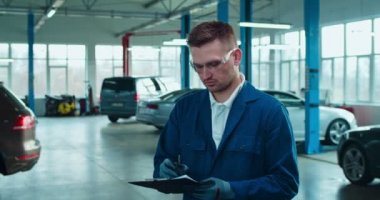 Formsuz ve gözlüklü yakışıklı beyaz adam koridorda arabalarla duruyor ve dosyaları dolduruyor. Garajda gülümseyen mutlu adamın portresi. Otomobil bakım kavramı.