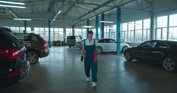 Biała młoda, atrakcyjna kobieta naprawiająca samochody w mundurze, goglach, kapeluszu i z kluczem zbliżającym się do kamery w dużym garażu. Portret bardzo szczęśliwej dziewczyny mechanik uśmiechnięty. — Wideo stockowe
