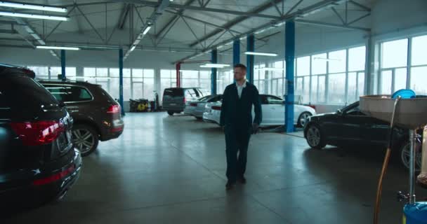 Blanc jeune homme beau travailleur de réparation de voitures en uniforme, lunettes et avec clé à approcher de la caméra dans un grand garage. Portrait de mec cool mécanicien souriant joyeusement et posant . — Video