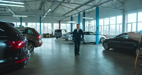 Retrato do homem legal bonito jovem caucasiano auto reparador trabalhador em uniforme, óculos e com chave aproximando-se da câmera no salão de grandes carros. Cara mecânica de trabalho posando com sorriso feliz . — Vídeo de Stock