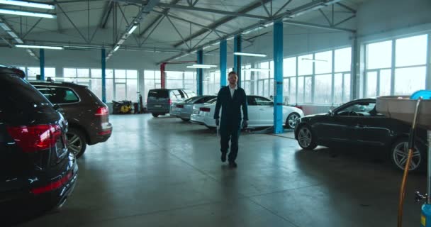 Portrait shot of Caucasian young handsome cool man auto mechanic in overalls, goggles and with wrench coming close to camera in big cars garage. Guy working in vehicles repairment with cheerful smile. — Stock Video