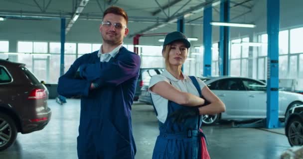 Retrato de mulher branca bonita e homem bonito. Dois mecânicos de automóveis em uniformes de pé no salão com carros. Menina e cara sorrindo para câmera alegremente na garagem. Conceito de manutenção de automóveis . — Vídeo de Stock