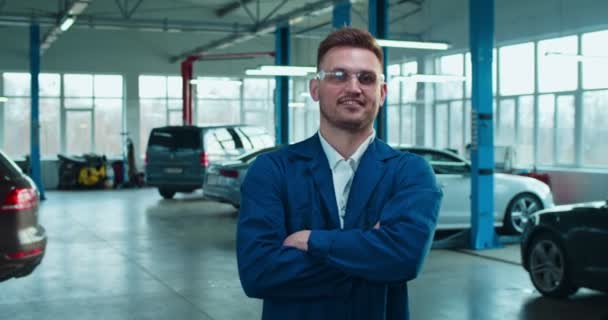 Portrait of handsome Caucasian man auto mechanic in unform and goggles standing in hall with cars. Happy guy smiling to camera cheerfully in garage. Automobile maintenance concept. — Stock Video