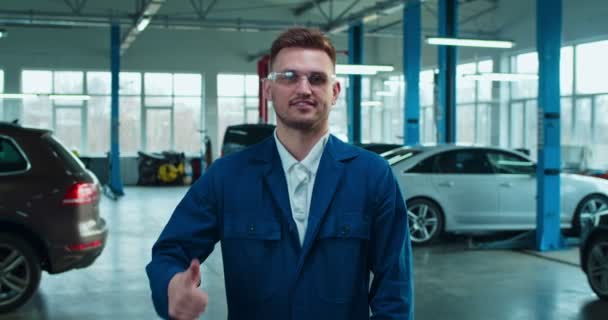 Retrato de joven caucásico hombre guapo mecánico de automóviles en gafas y uniforme sonriendo a la cámara y dando pulgar hacia arriba en el garaje. Joven chico alegre de pie en el servicio de coches . — Vídeo de stock