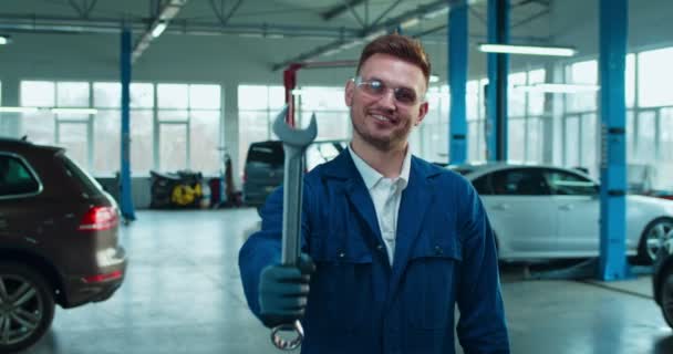 カメラに笑みを浮かべてガレージでレンチを示す幸せな陽気な白人ハンサムな男の自動車整備士の肖像画。若いです男労働者で全体とゴーグル保持ツールで車のサービス. — ストック動画
