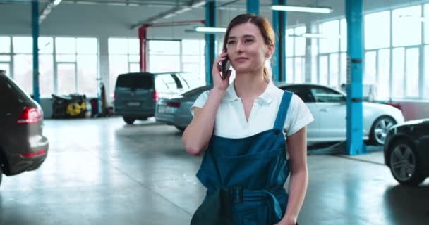 Bella donna caucasica meccanico auto in uniforme parlando sul telefono cellulare. Bella ragazza sorridente e parlando al cellulare in garage auto. Concetto di riparazione di automobili. conversazione telefonica di femmina . — Video Stock