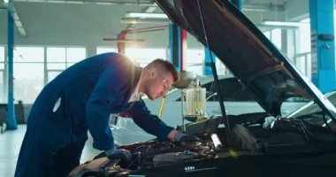 Beyaz bir erkek, genç araba tamircisi açık arabada duruyor ve büyük bir garajda motor tamir ediyor. Üniformalı ve gözlüklü bir adam otomobil tamirhanesinde çalışıyor. Taşıma hizmeti kavramı.