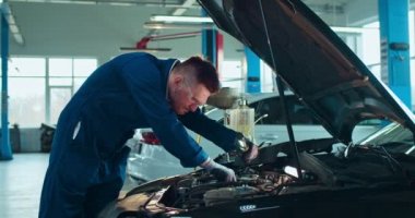 Beyaz tenli genç erkek oto tamircisi açık arabada duruyor ve büyük garajda ışıkla motoru tamir ediyor. Üniformalı ve gözlüklü bir adam otomobil tamirhanesinde çalışıyor. Araba servisi kavramı.