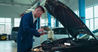 Beyaz erkek, genç araba tamircisi açık arabada duruyor ve tablet cihazıyla motoru tamir ediyor. Üniformalı ve gözlüklü bir adam otomobil servisinde çalışıyor. Bilgisayarı tıklatıyor ve ipuçlarını izliyor..