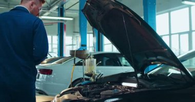 Üniformalı ve gözlüklü beyaz bir genç arabayla motoru açmaya ve bazı belgeleri kalemle doldurmaya geliyor. Otomobil servis yazma ve otomobil koşullarını not eden erkek işçi.