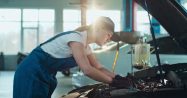 Beyaz kadın otomobil mühendisi açık arabada duruyor ve büyük atölyede motorunu tamir ediyor. Üniformalı, gözlüklü, kapalı otoparkta çalışan bir kadın. Taşıma hizmeti kavramı.