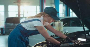 Beyaz kadın otomobil mühendisi açık arabada duruyor ve büyük atölyede motorunu tamir ediyor. Yardım eden ve öğreten adam. Kadın ve erkek işçiler otomobil servis garajında kapalı alanda üniforma giyiyorlar..