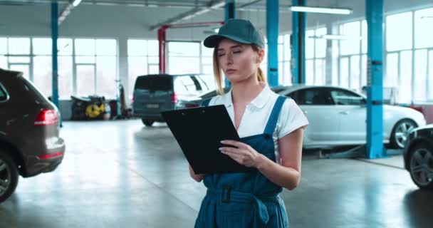 Vacker vit kvinna auto mekaniker i form och hatt står i hallen med bilar och fylla i dokument i mapp. Ung söt flicka skriver på papper i garaget. Underhållskoncept för bilar. — Stockvideo