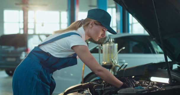 Vrouwelijke blanke jonge automonteur staat bij open auto en repareert motor in garage, mannelijke collega komt helpen. Een vrouw en een man die binnen in een garage werken. Dienstconcept. — Stockvideo