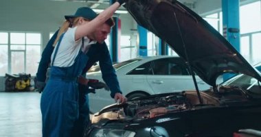 Beyaz kadın otomobil tamircisi araba motoru açıyor ve erkek meslektaşına büyük garajda kırılma sorunu gösteriyor. Erkek ve kadın mühendisler otomobil üzerinde birlikte çalışıyorlar..