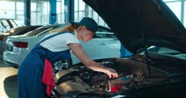 Beyaz kadın araba tamircisi açık arabada duruyor ve büyük garajda motor tamir ediyor. Üniformalı kadın portresi otomobil tamirinde çalışıyor ve kameraya neşeyle gülümsüyor..