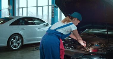 Beyaz kadın araba tamircisinin portresi açık arabada duruyor ve büyük garajda motor tamir ediyor. Üniformalı sarışın kadın otomobil tamirinde çalışıyor ve kameraya neşeyle gülümsüyor..