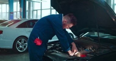 Beyaz bir erkek, genç araba tamircisi açık arabada duruyor ve büyük bir garajda motor tamir ediyor. Üniformalı bir adamın portresi otomobil tamirinde çalışıyor ve kameraya neşeyle gülümsüyor..