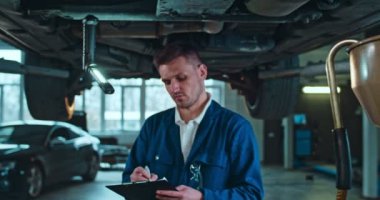 Beyaz tenli yakışıklı erkek otomobil tamircisinin portresi. Kameraya bakıyor ve dosyayla gülümsüyor. Garajda ellerinde belge var. Teknik inceleme sırasında arabanın altında duran adam.