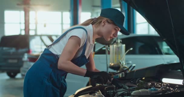 Caucasienne jeune mécanicien automobile femme debout à la voiture ouverte et réparer le moteur avec lampe de poche dans un grand garage. Fille en uniforme et chapeau de travail à l'intérieur salon de réparation automobile. Concept de service de voitures . — Video