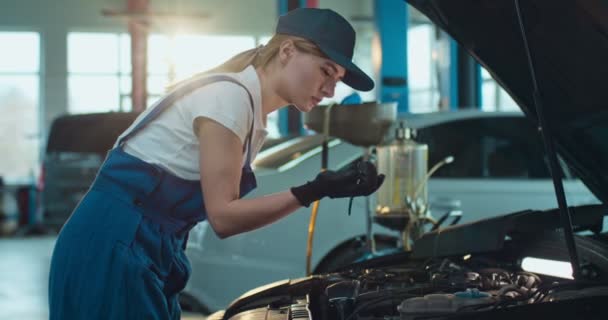 Mujer hermosa joven caucásica en uniforme y sombrero con linterna en la mano y en busca de ruptura en el motor del coche en el garaje. Trabajadora de autoservicio reparando roturas en automóvil en taller . — Vídeos de Stock