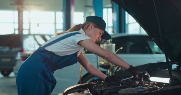 Kobieta Kaukaski młody mechanik samochodowy stoi przy otwartym samochodzie i naprawia silnik w dużym garażu. Kobieta w mundurze i kapelusz serwisowania pojazdu w salonie naprawy samochodów wewnątrz. Koncepcja usług transportowych — Wideo stockowe