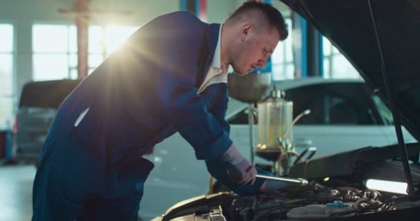 Mężczyzna biały młody mechanik samochodowy stojący przy otwartym samochodzie i naprawiający przerwy w dużym garażu. Mężczyzna w mundurze i gogle pracujący w salonie samochodowym. Koncepcja usług transportowych. — Wideo stockowe