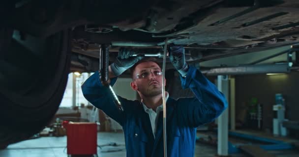 Caucasiano jovem bonito homem de uniforme e óculos de pé sob o carro e torcendo alguns detalhes com chave na garagem. Mecânico masculino trabalhando no salão de serviço de automóveis e pausas de reparação . — Vídeo de Stock